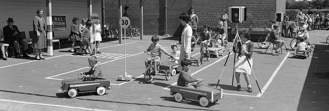 Black and white photo of school children riding toy cars to learn road safety circa 1950s.