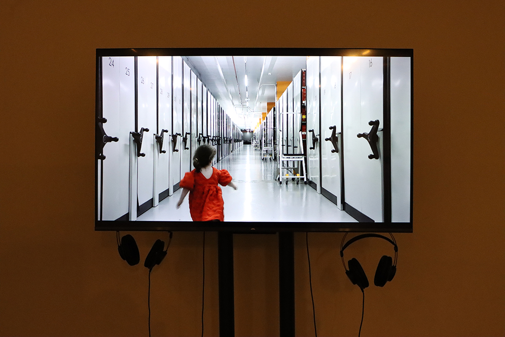 Rewind Forward exhibition. A video screen of a small girl in a red dress running through the archival repository. Underneath the screen are 2 sets of headphones.