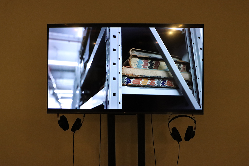 Photo of a screen with two headphones hanging beneath it. The image on the screen is old books on a metal shelf.