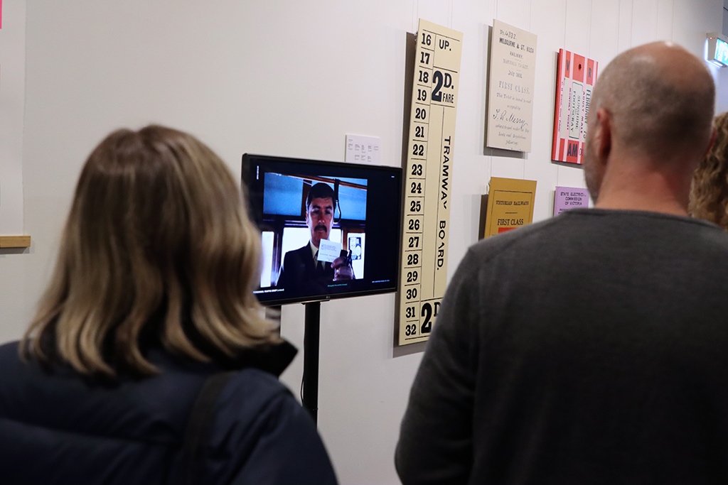 Rewind Forward opening night. People are viewing a slideshow on a screen. Behind it are paintings on a wall.