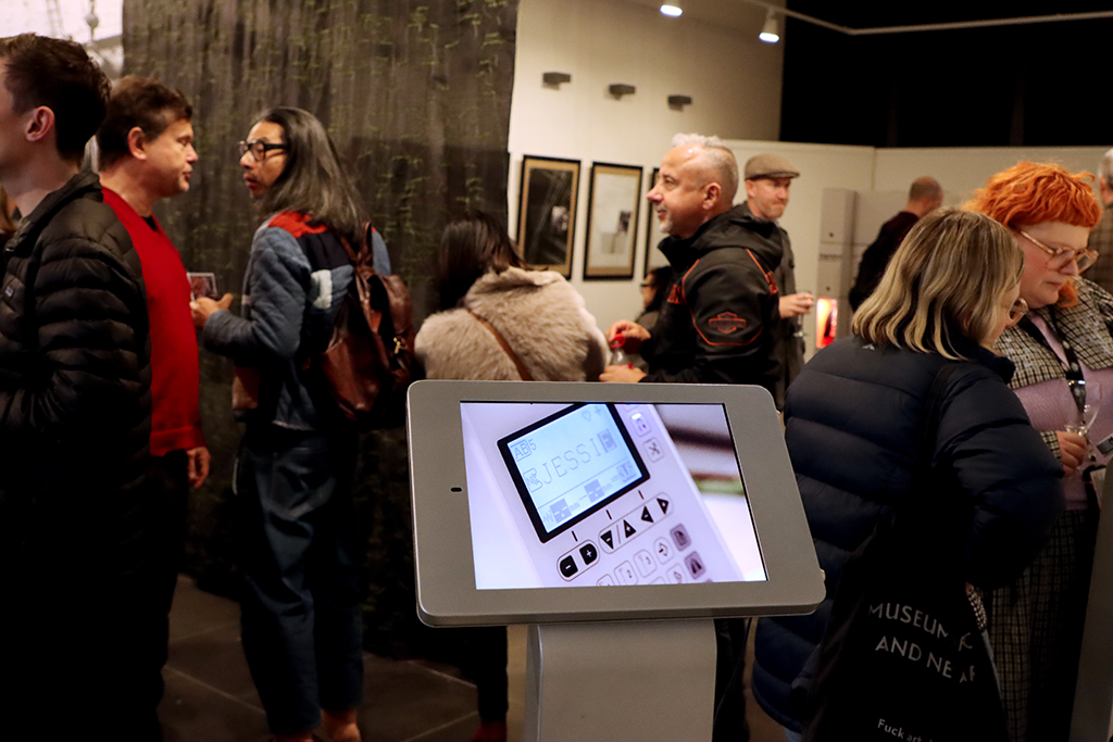 An iPad on a stand is in the foreground Behind are people walking through the gallery. In the background is an artwork made from a large vertical piece of black silk.