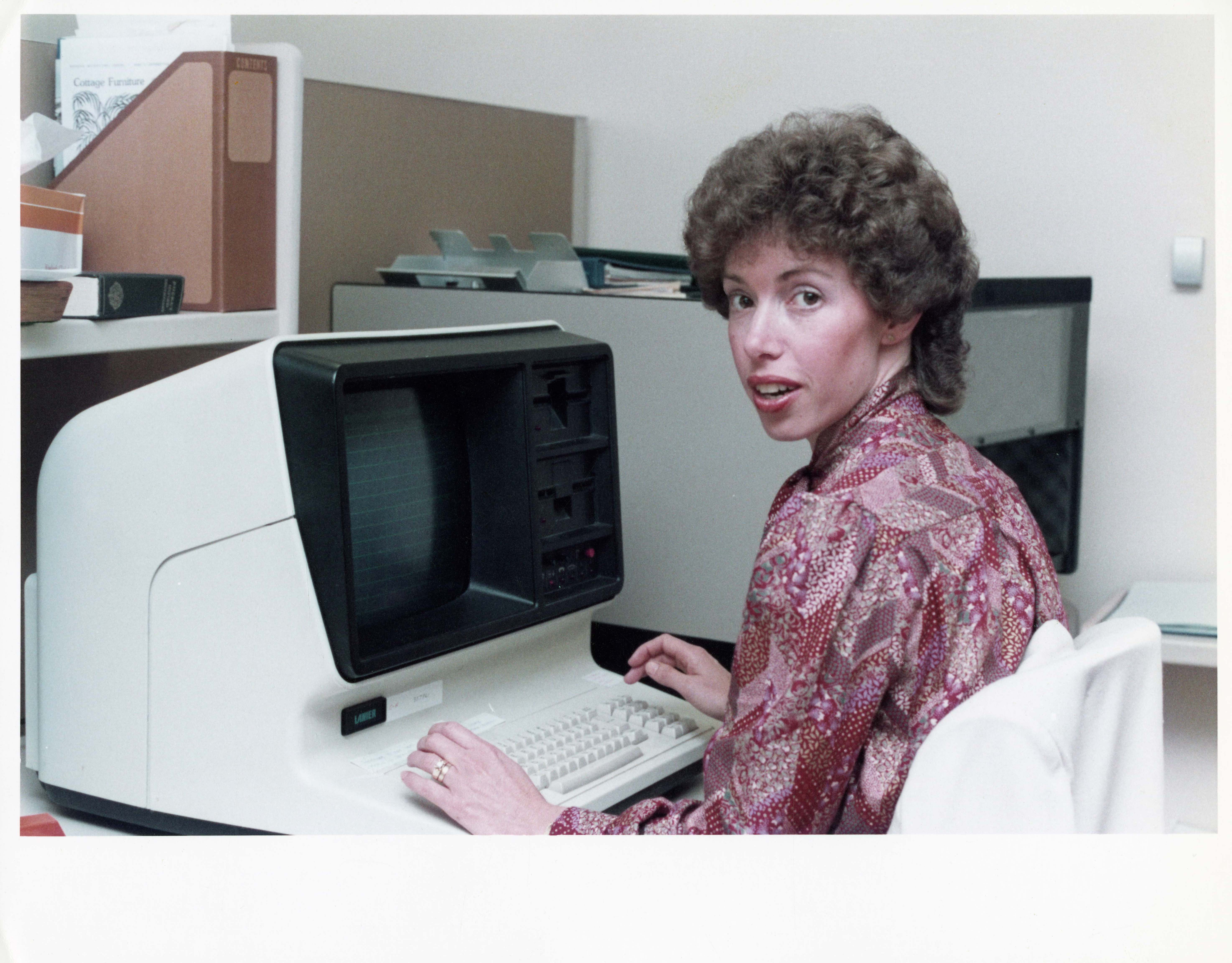 colour photo of a woman with an 80s perm typing at a large computer