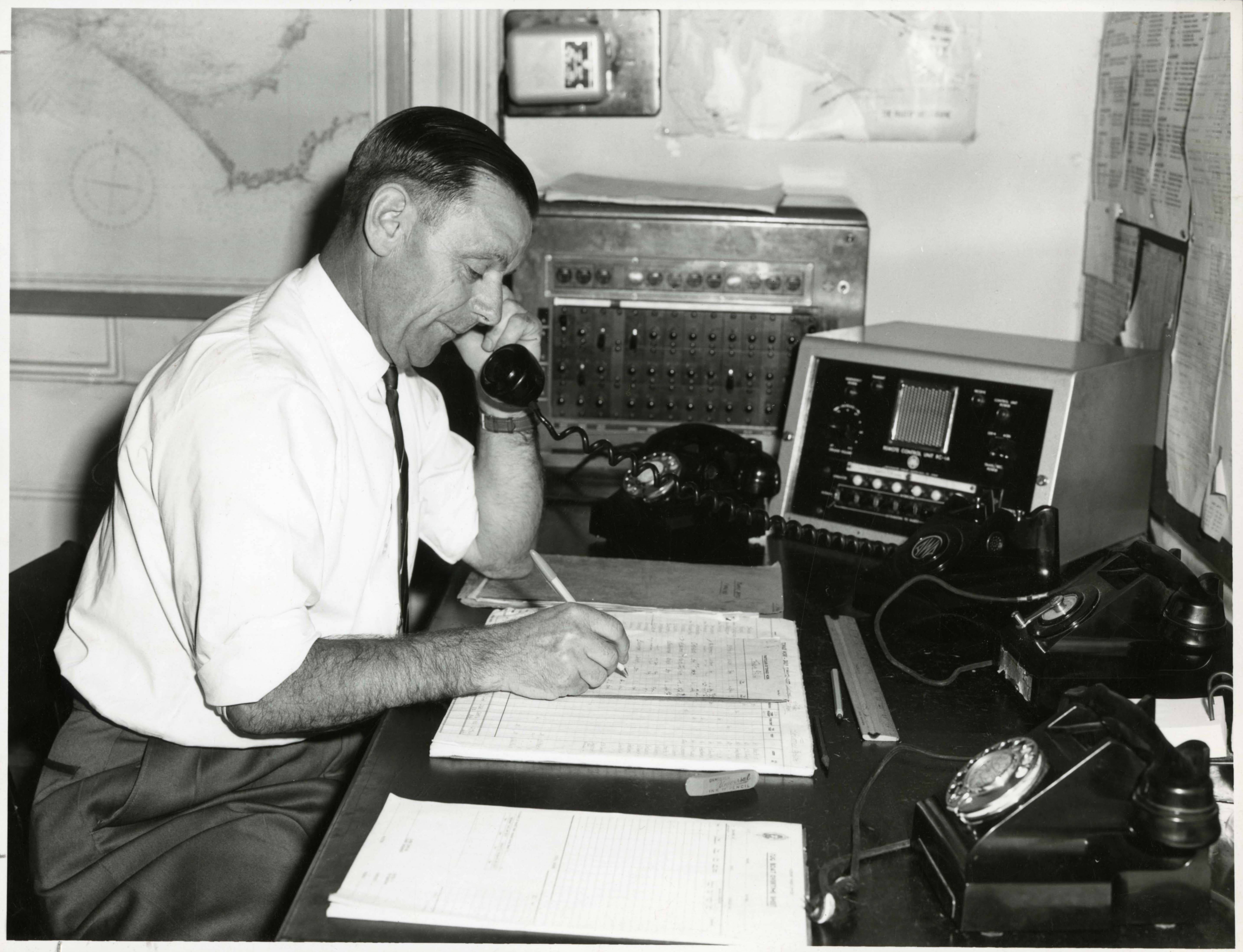 black and white photo man at desk with three phones, he is on one