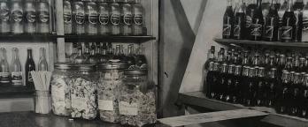 black and white photo of bottles stacked in a shop with lollie jars on the counter