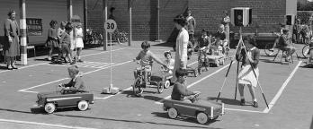 Black and white photo of school children riding toy cars to learn road safety circa 1950s.