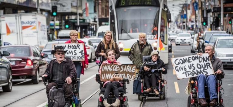 Colour photograph of a group of people in front of a tram on a Melbourne street, protesting the lack of tram accessibility. Four people are using wheelchairs and three people walk behind them.