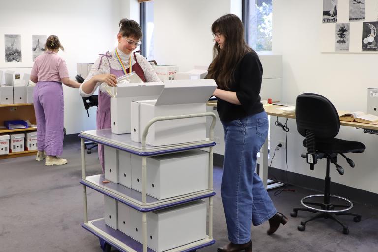 Three women working in an office with a trolley loaded with boxes of archival records